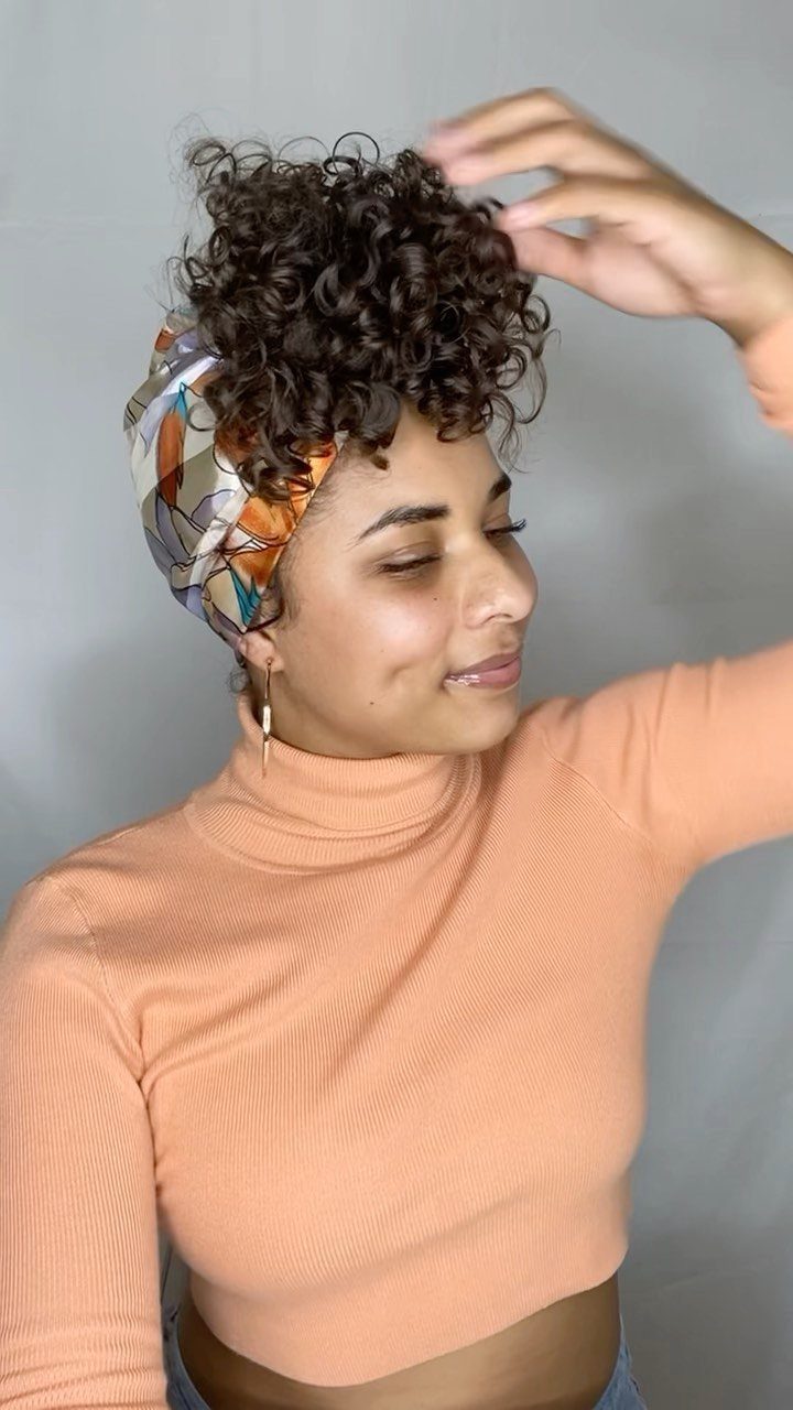 Person with short dark curls gathered high, touching their hair while wearing a patterned scarf