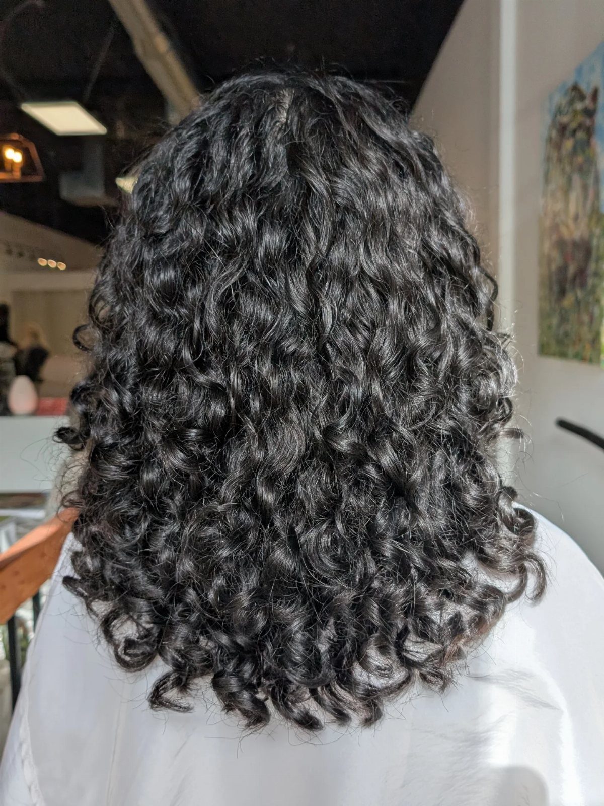 Dark shoulder-length curls show a rounded lob shape from behind in a salon.
