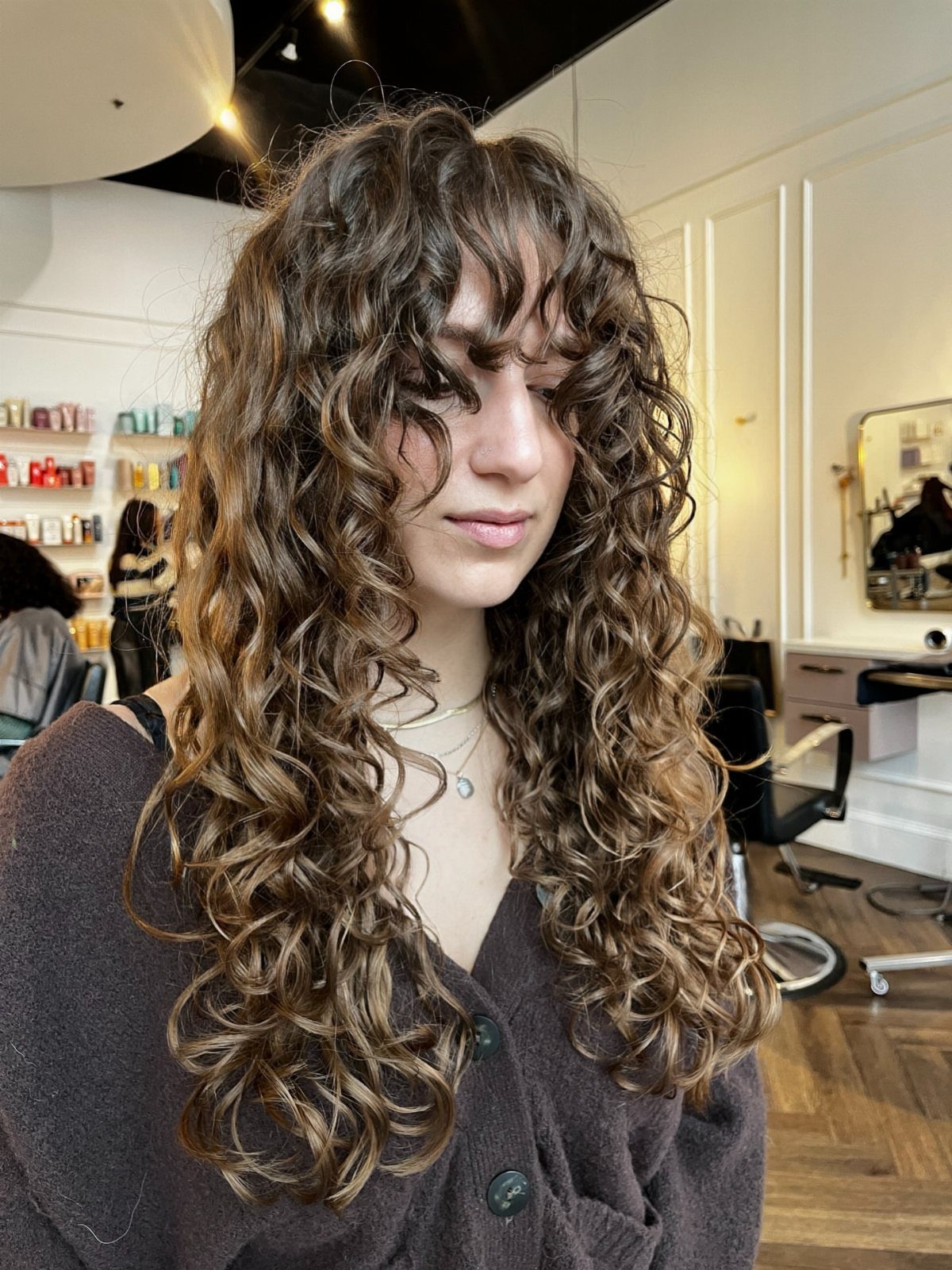 Long curly hair with bangs falls in rounded layers around a seated salon client