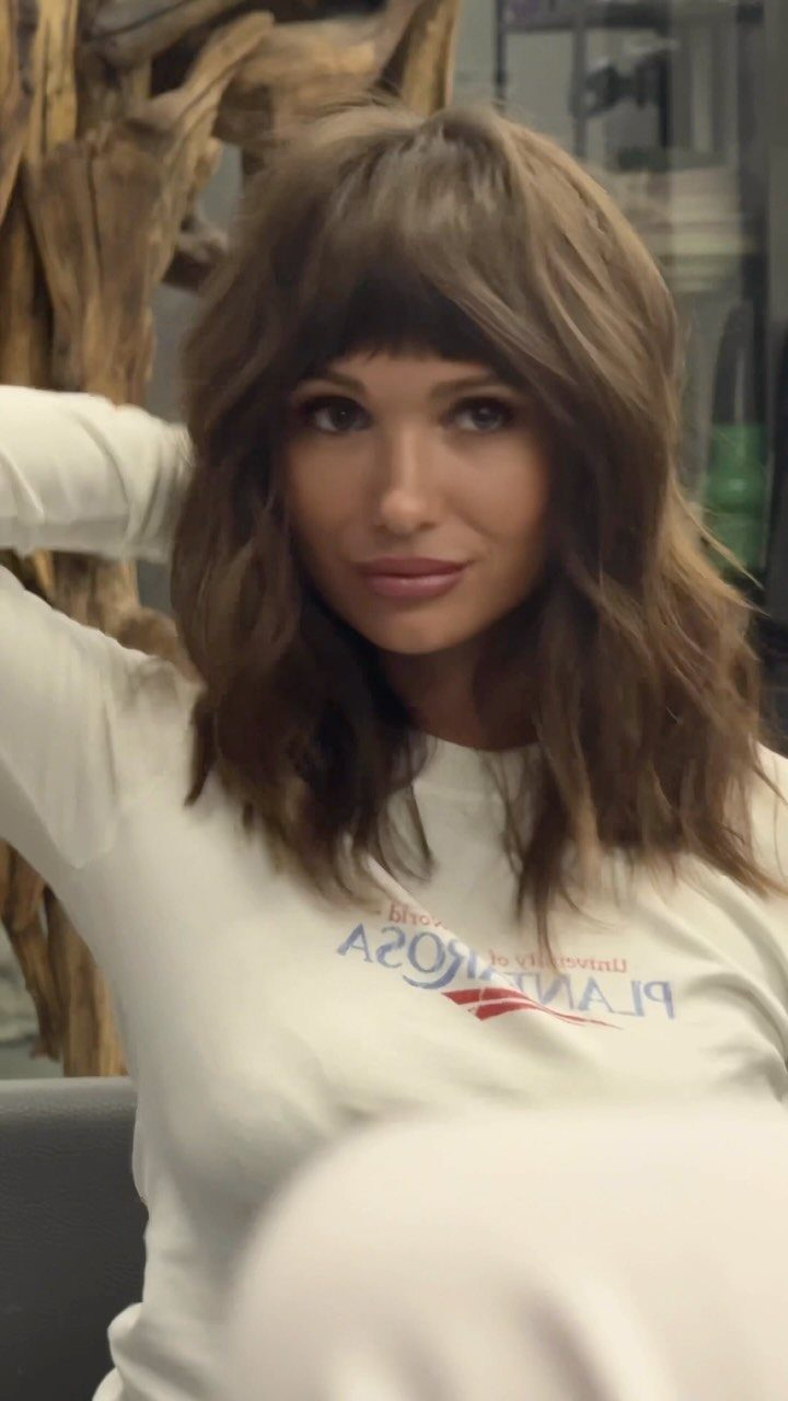 Woman with long wavy brown hair and softly feathered bangs in a white sweatshirt