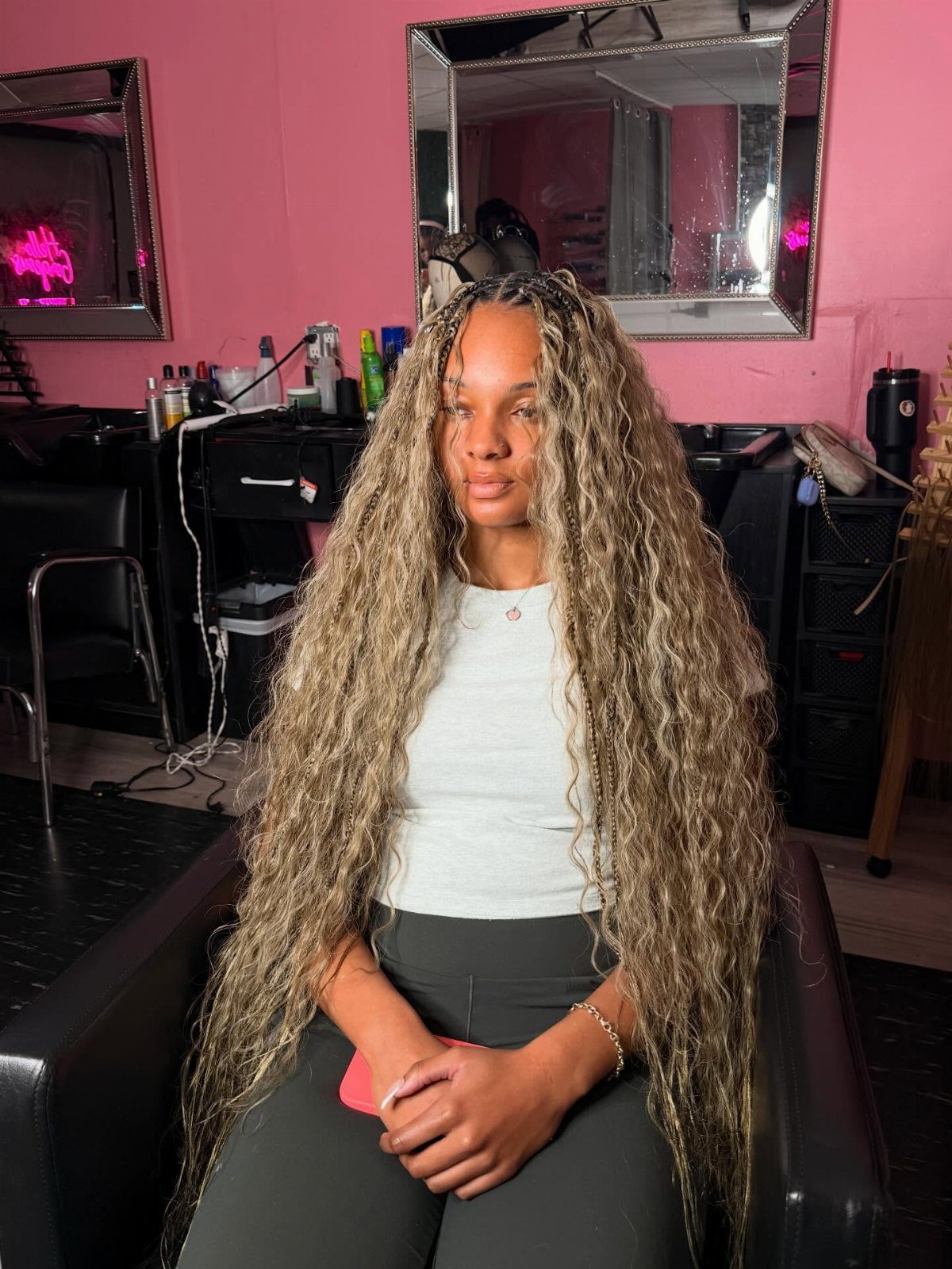 Woman with long blonde boho braids and dark roots seated in a pink salon