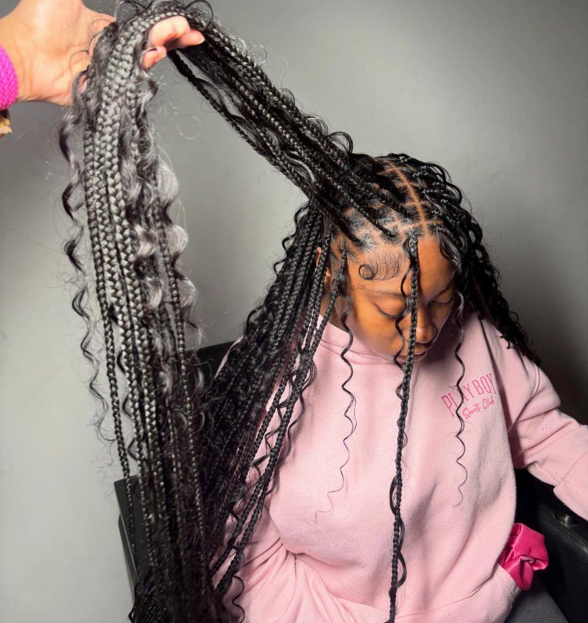 Long black knotless braids with loose curls held upward over a pink sweatshirt