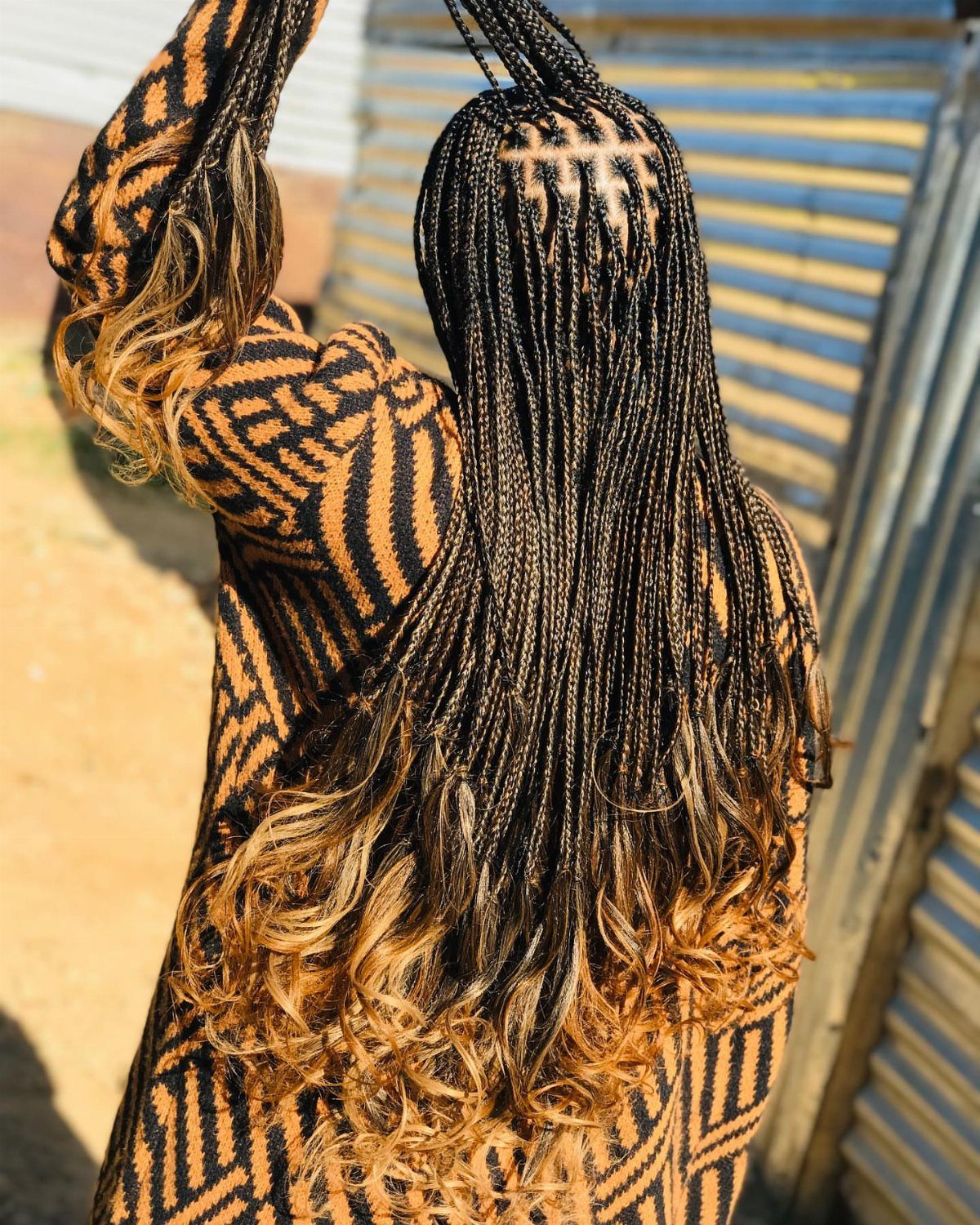 Long black braids with caramel curly ends cascade down a patterned outfit