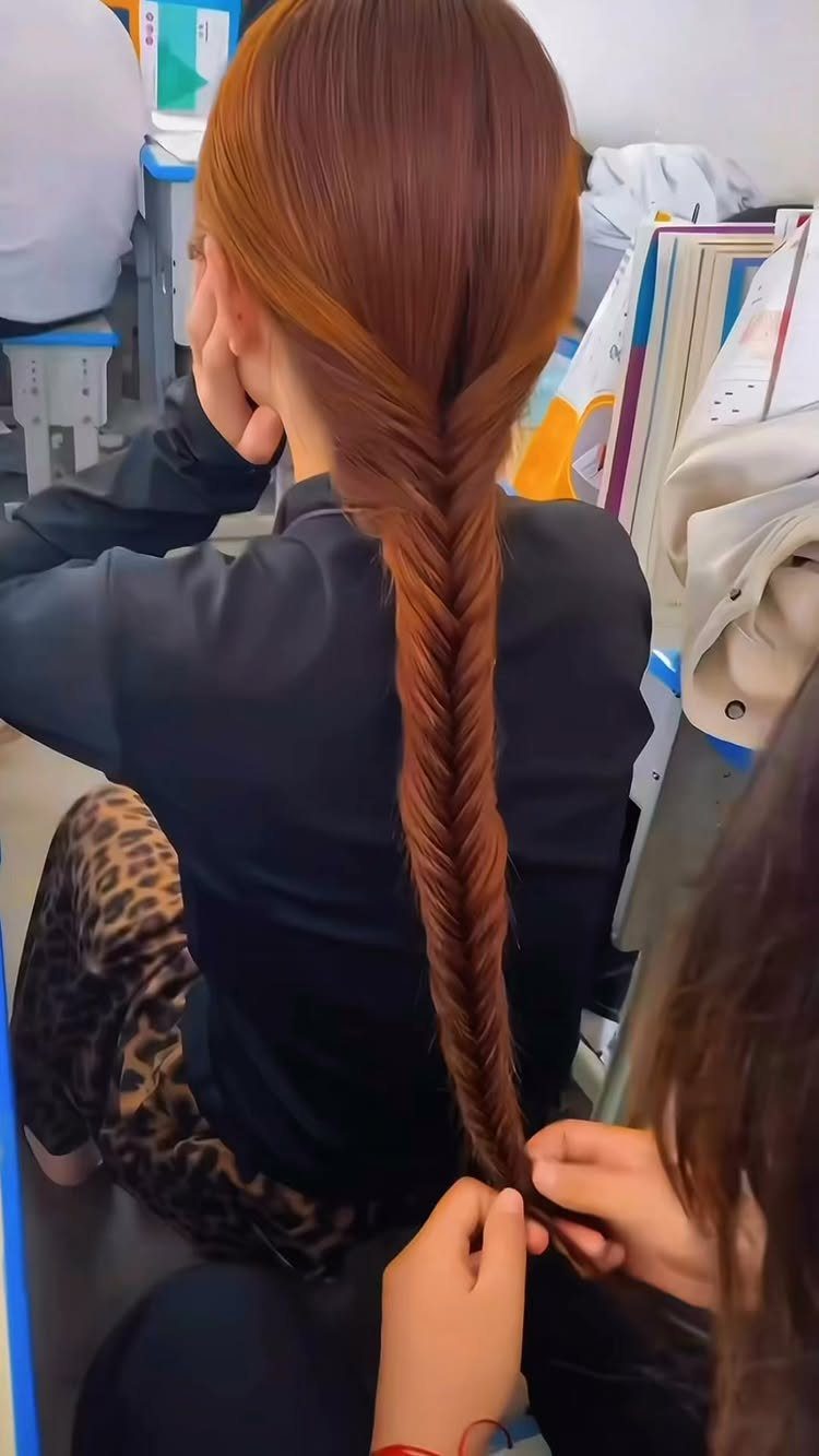 Auburn-haired person receiving a long, tight braided ponytail hairstyle