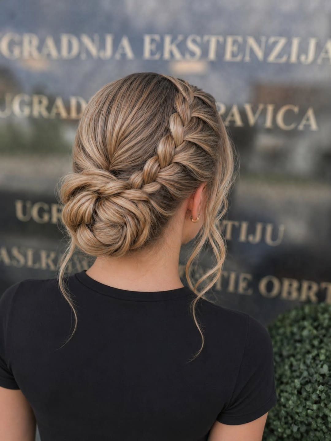 Woman showing a braided low side bun with loose tendrils against a blurred sign