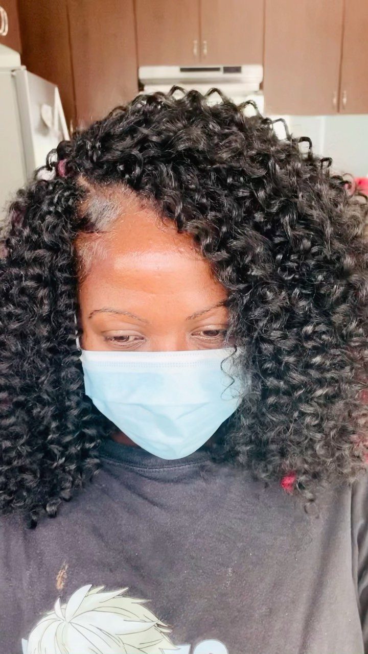 Side-parted black curly crochet braids frame a masked person’s face