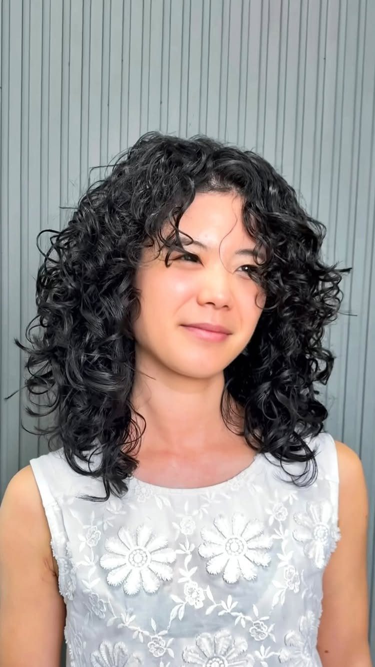 Person with shoulder-length layered black curls posing against a pale paneled wall