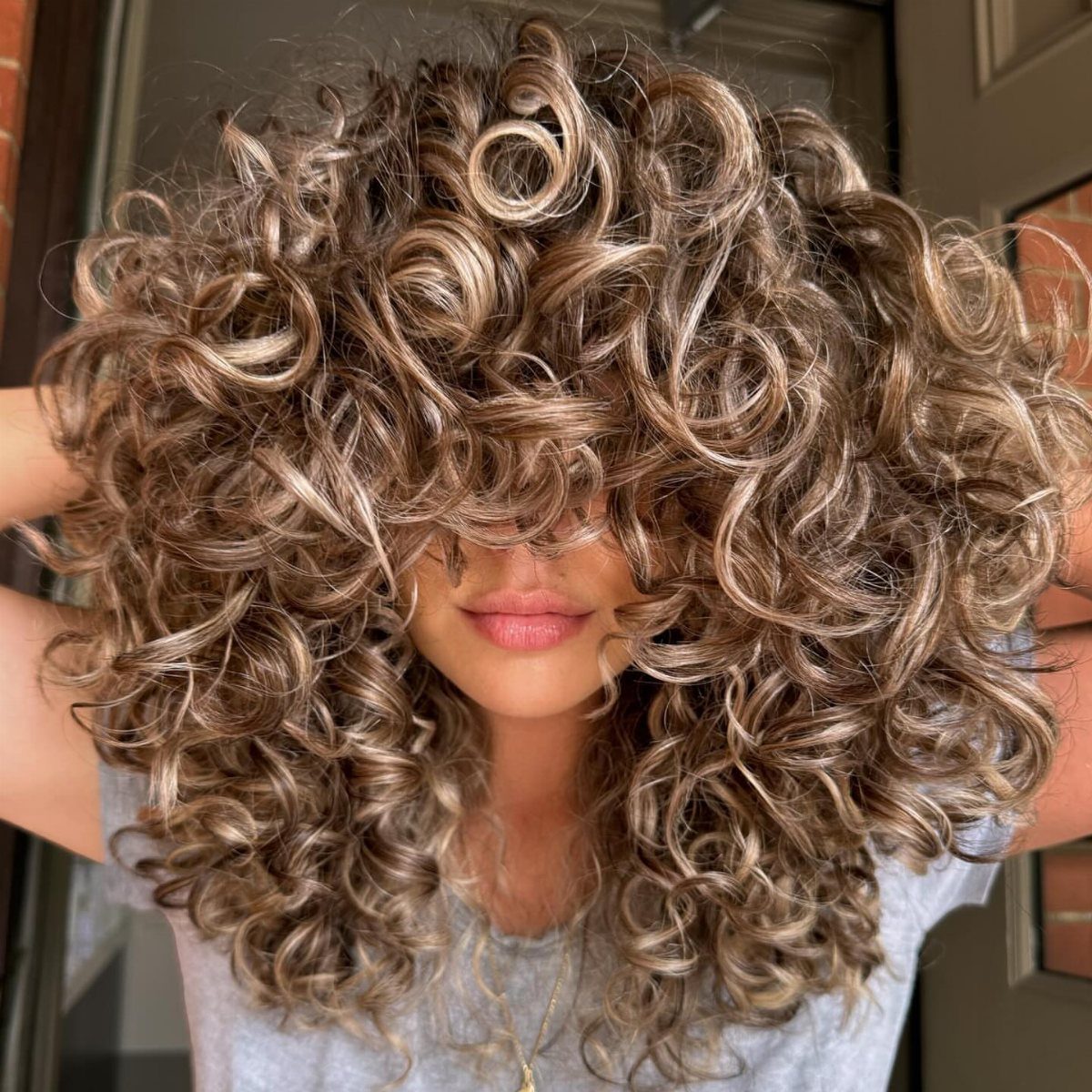 Voluminous caramel-highlighted curls frame a person's face in a doorway