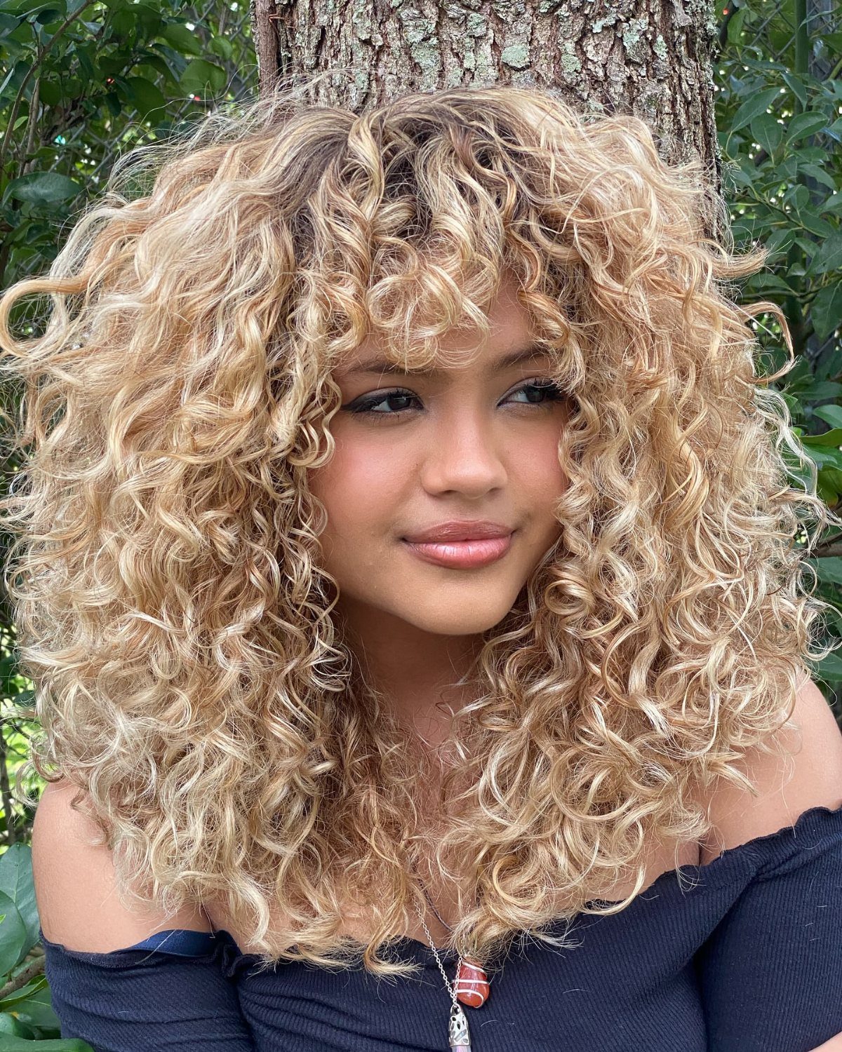 Shoulder-length golden curls frame a person posing in front of a tree