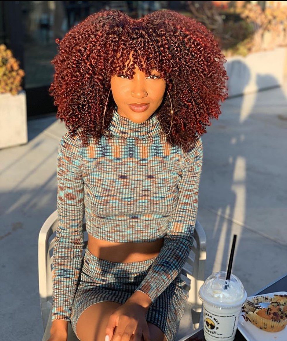 Woman with reddish-brown coils and short bangs seated outdoors beside a drink