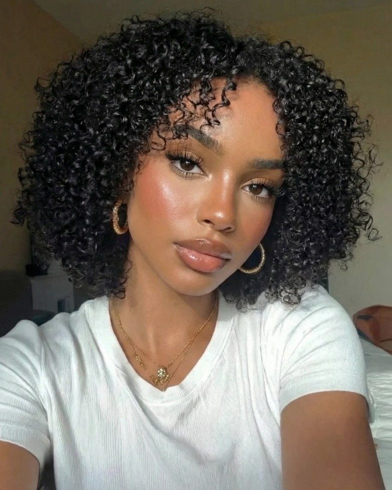 Person with dense black curls and short forehead-framing bangs posing in a white shirt