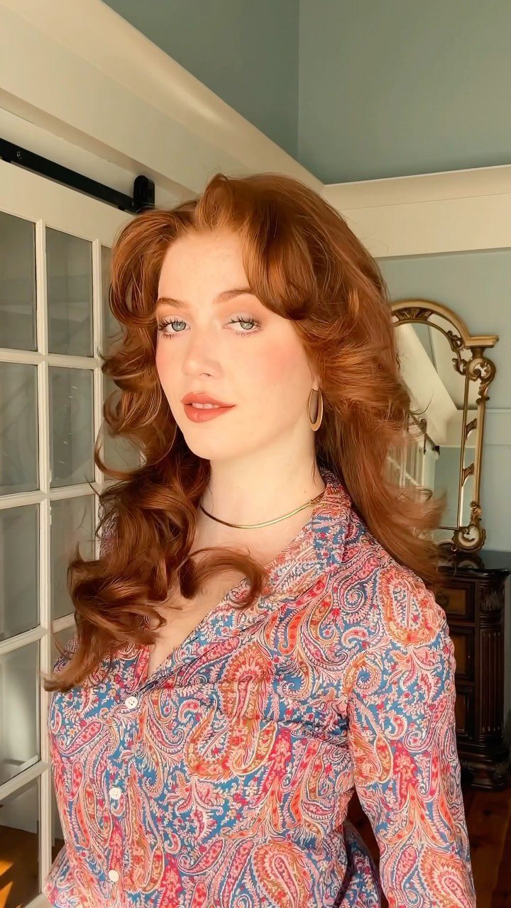 Woman with voluminous auburn curls and parted bangs wearing a colorful paisley blouse