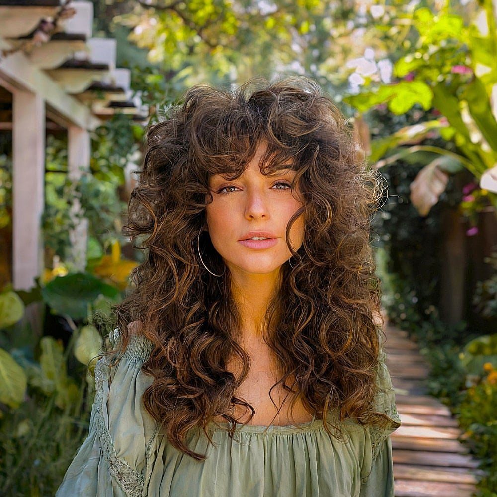 Woman with long layered curls and feather-light bangs in a leafy garden