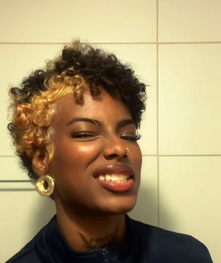 Short curly pixie with dark roots and golden-blond curls framing a smiling face