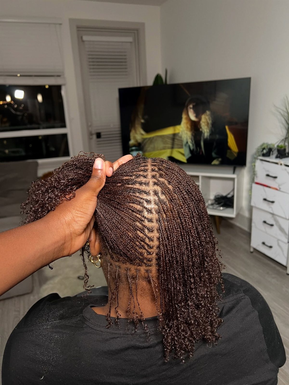 Micro braids with curly ends are parted by hand on the back of a person's head.