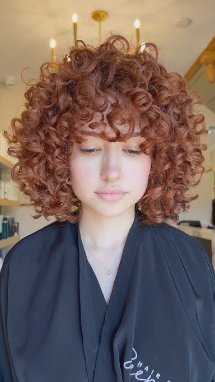 Short reddish-brown curls frame a person's face above a dark salon cape