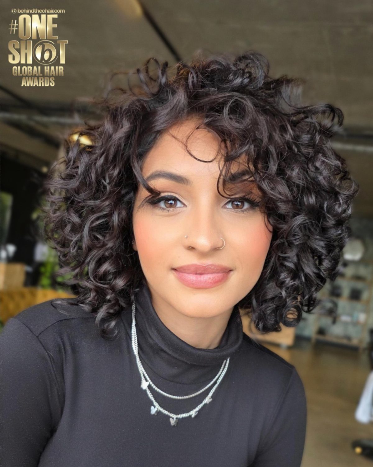 Woman with a voluminous chin-length curly bob, black top, and silver necklaces