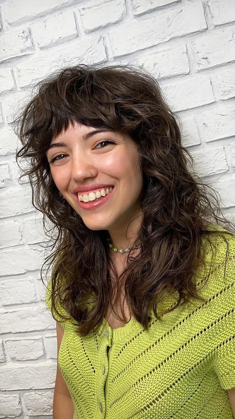 Woman with dark wavy long layers and short bangs against a white brick wall