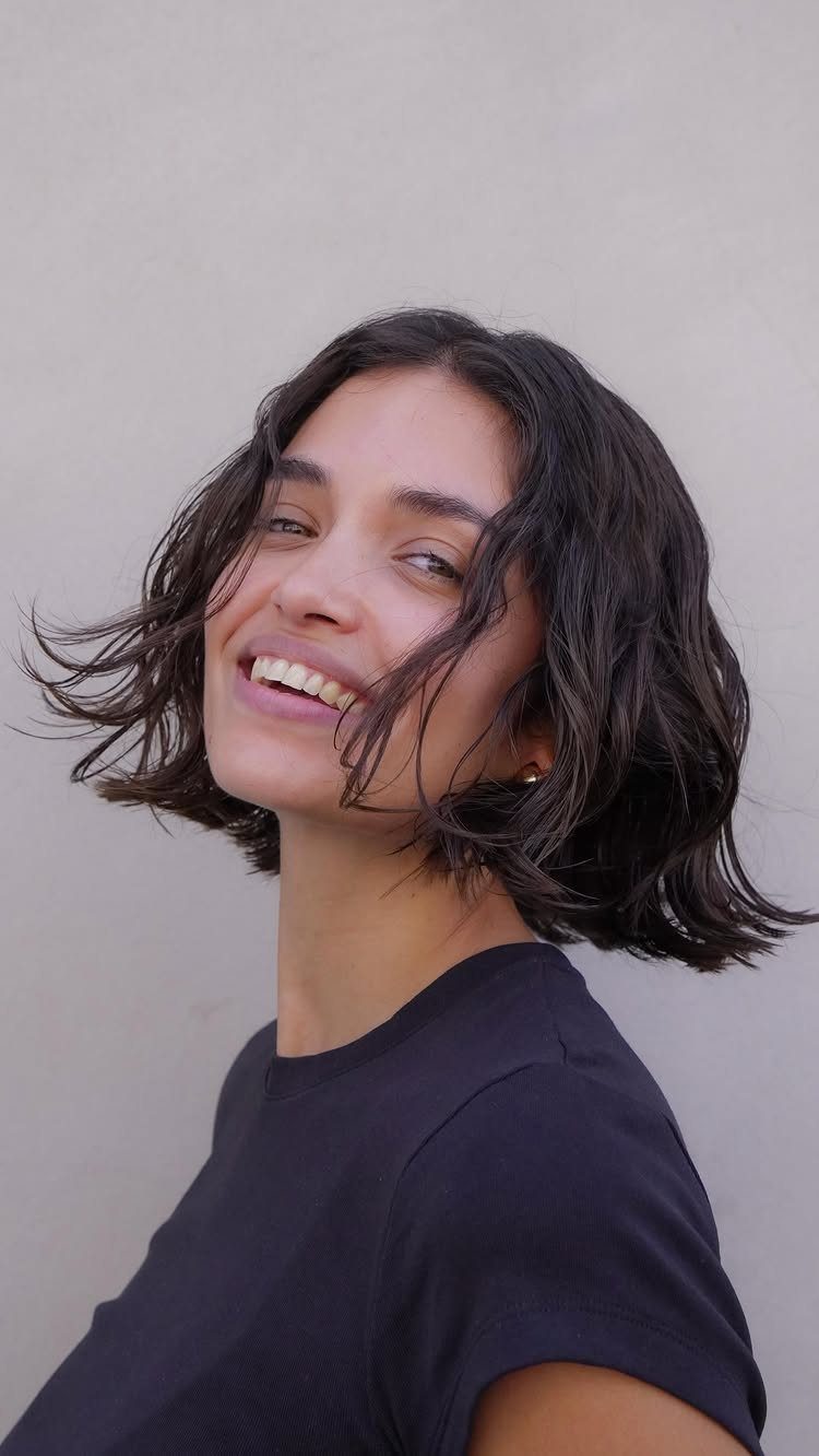 Short wavy bob frames a smiling person’s face against a plain wall.