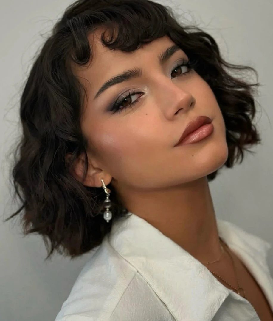 Woman with jaw-length dark curls and short curled bangs posing in a white top