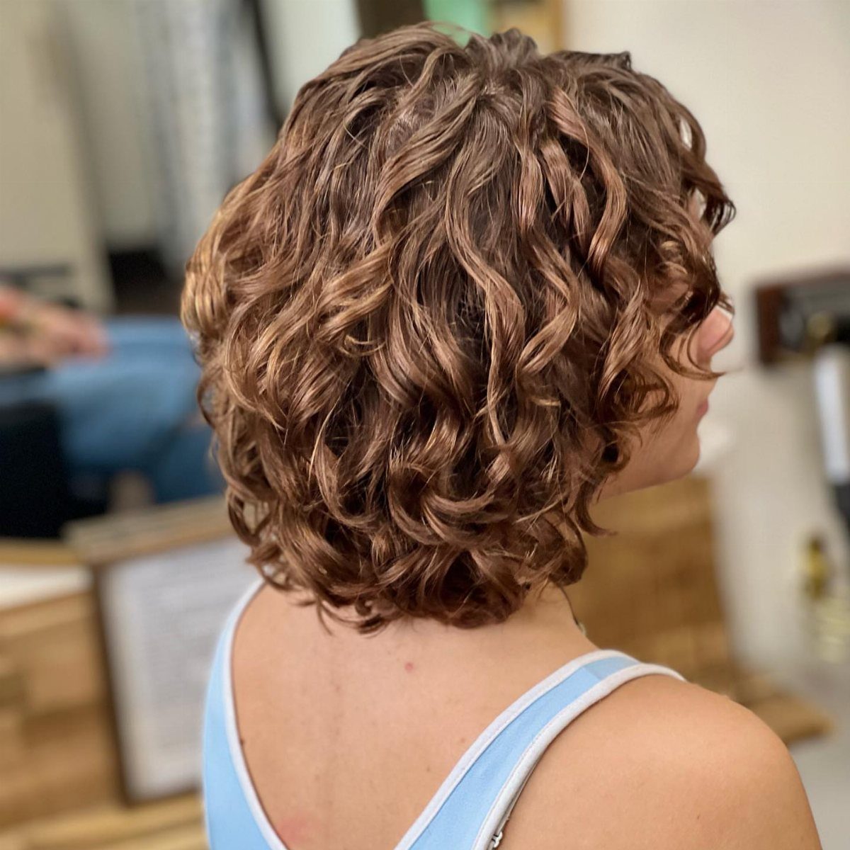 Short layered brown curls form a rounded, textured shape above a light blue top