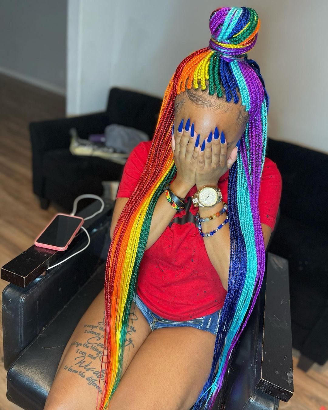 Rainbow braids hang long over a red shirt, with strands gathered into a high bun
