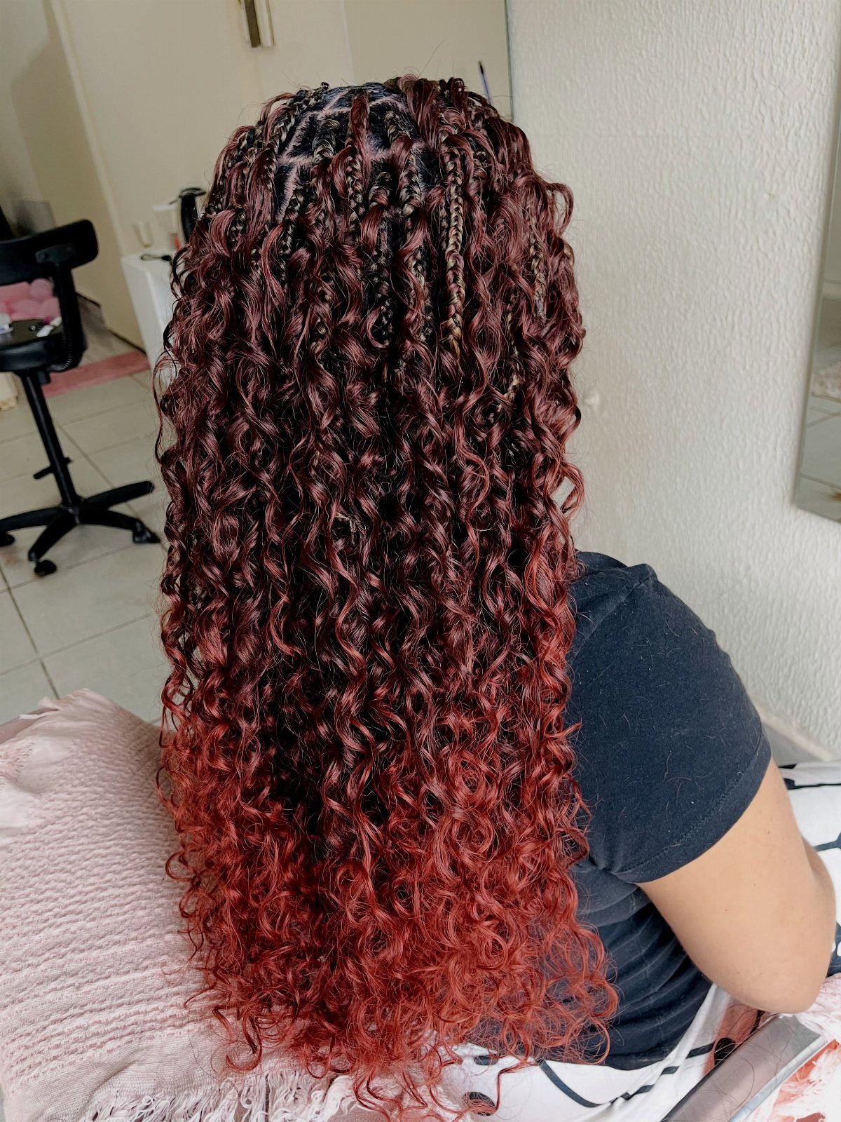 Long burgundy and red curly gypsy braids cascade down a seated person's back.