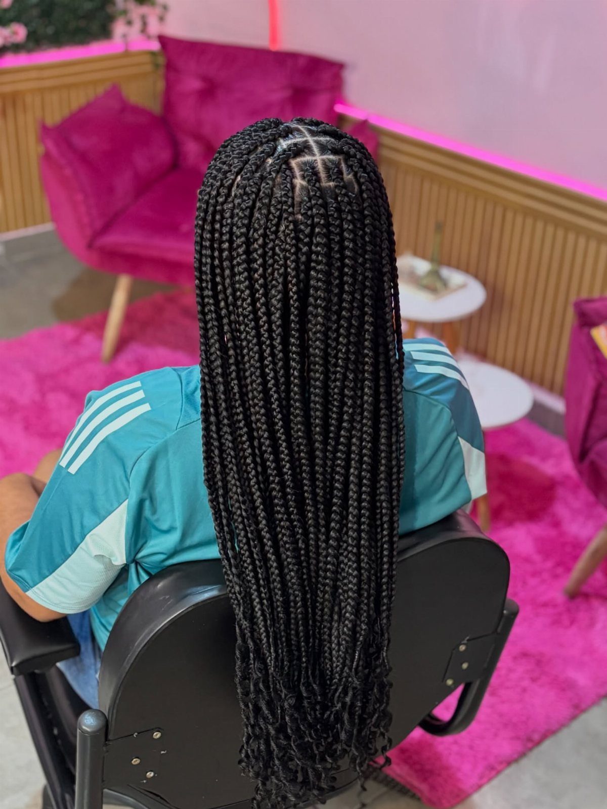 Long black box braids fall down a person's back in a bright pink salon.