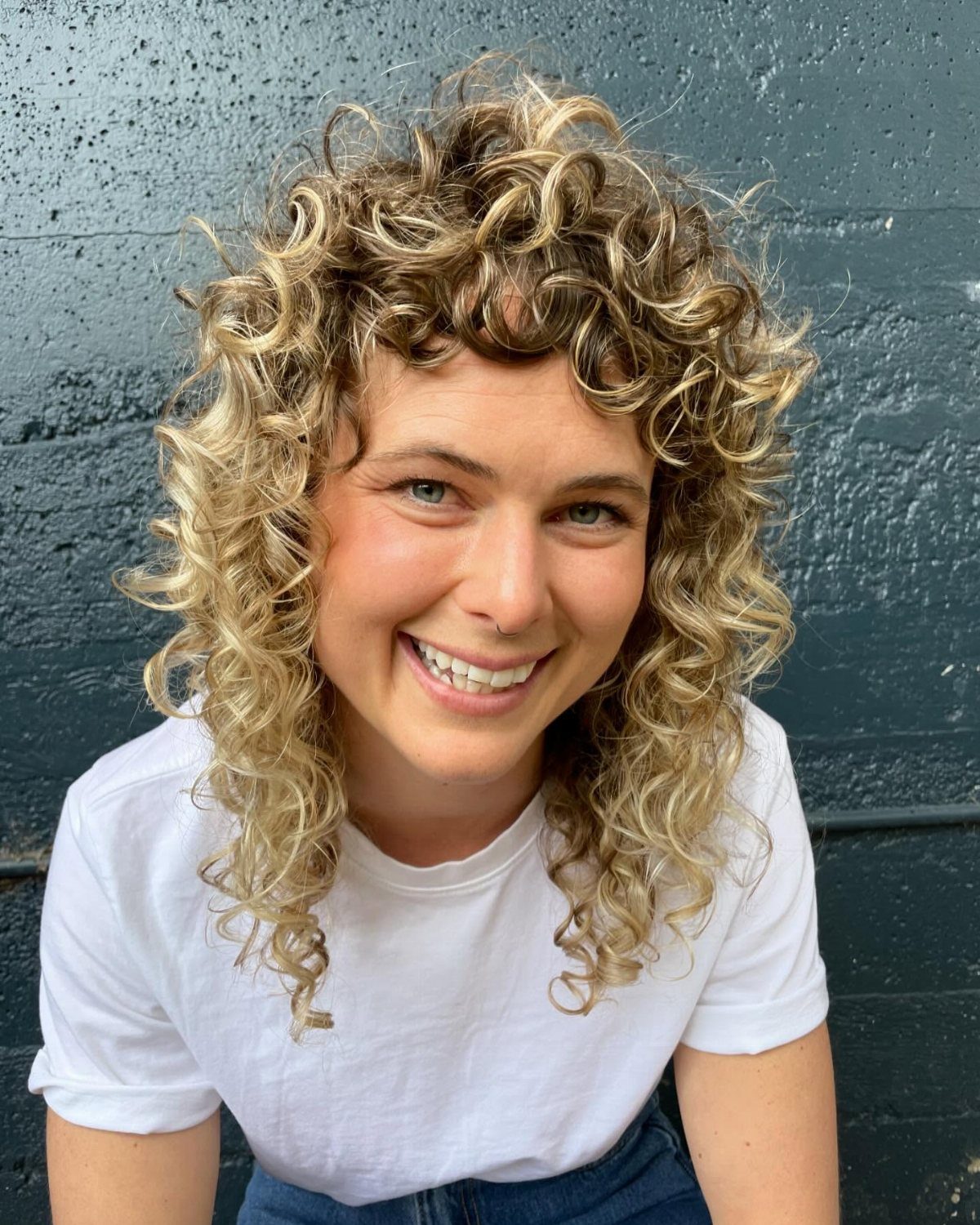 Smiling person with voluminous layered blonde curls against a dark wall