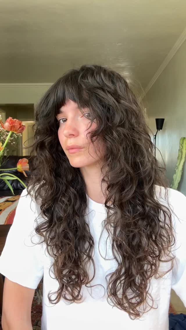 Long dark curls with a curly fringe frame a person wearing a white T-shirt