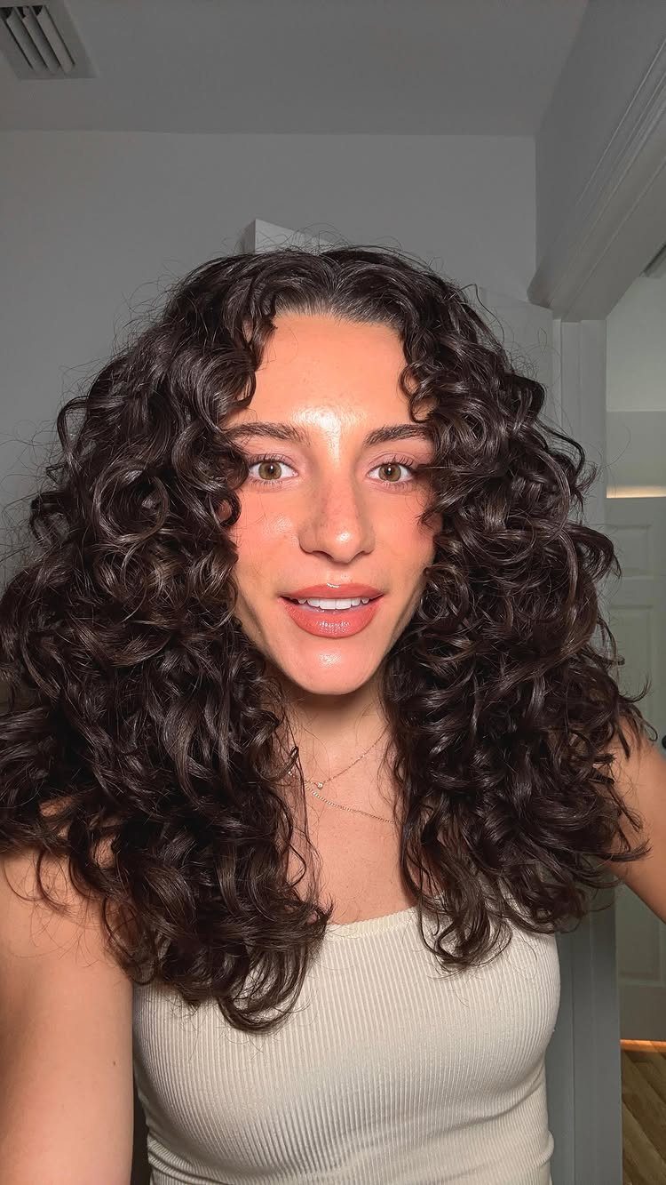 Long dark curls frame a person's face above a cream ribbed top