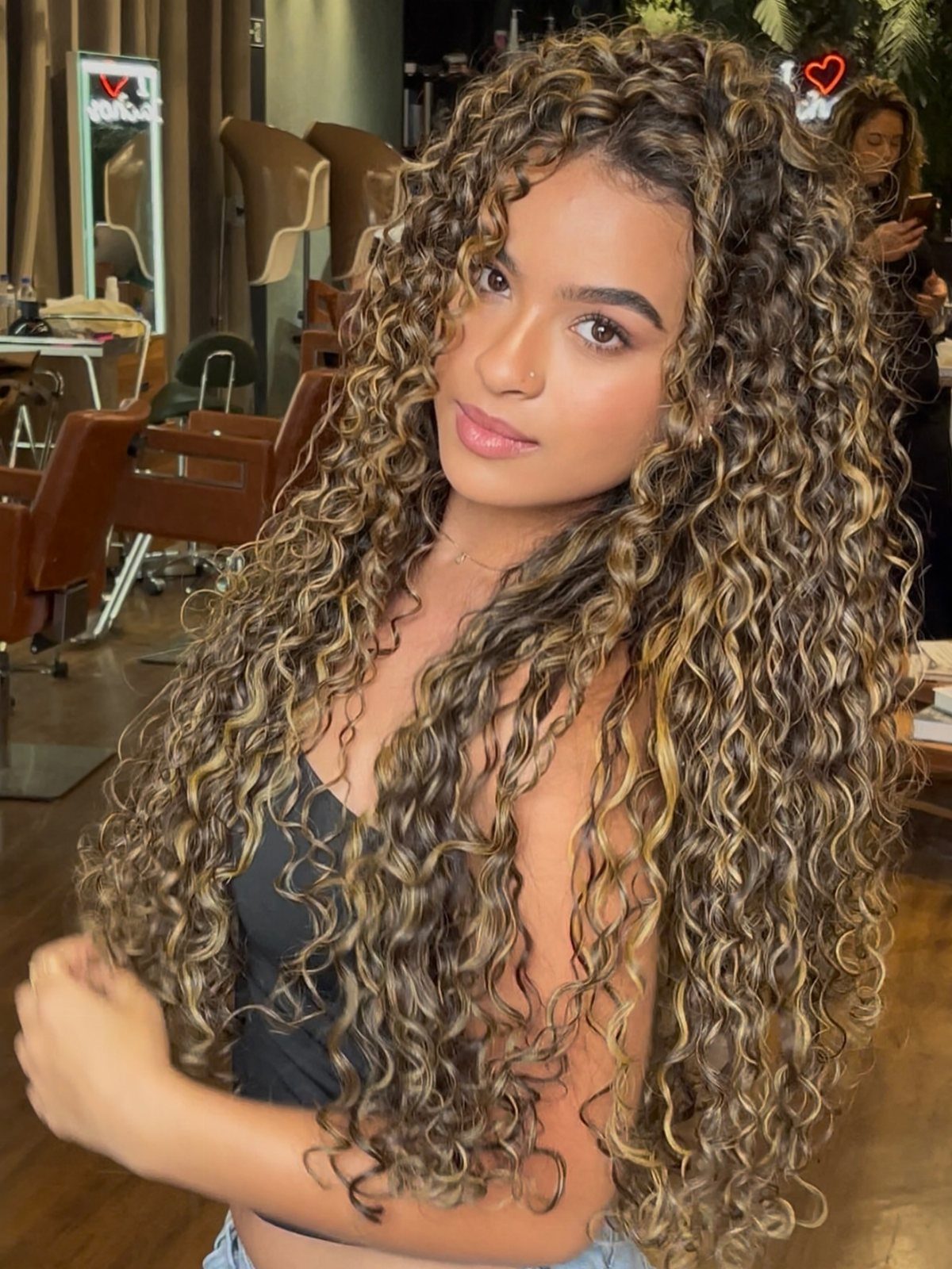 Woman with long dark curls and caramel highlights posing in a salon