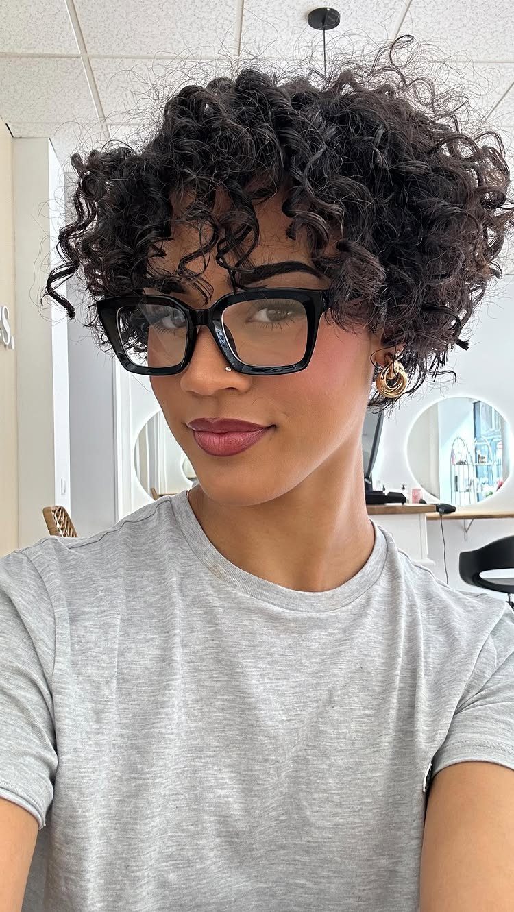 Rounded short dark curls frame a person wearing oversized black glasses