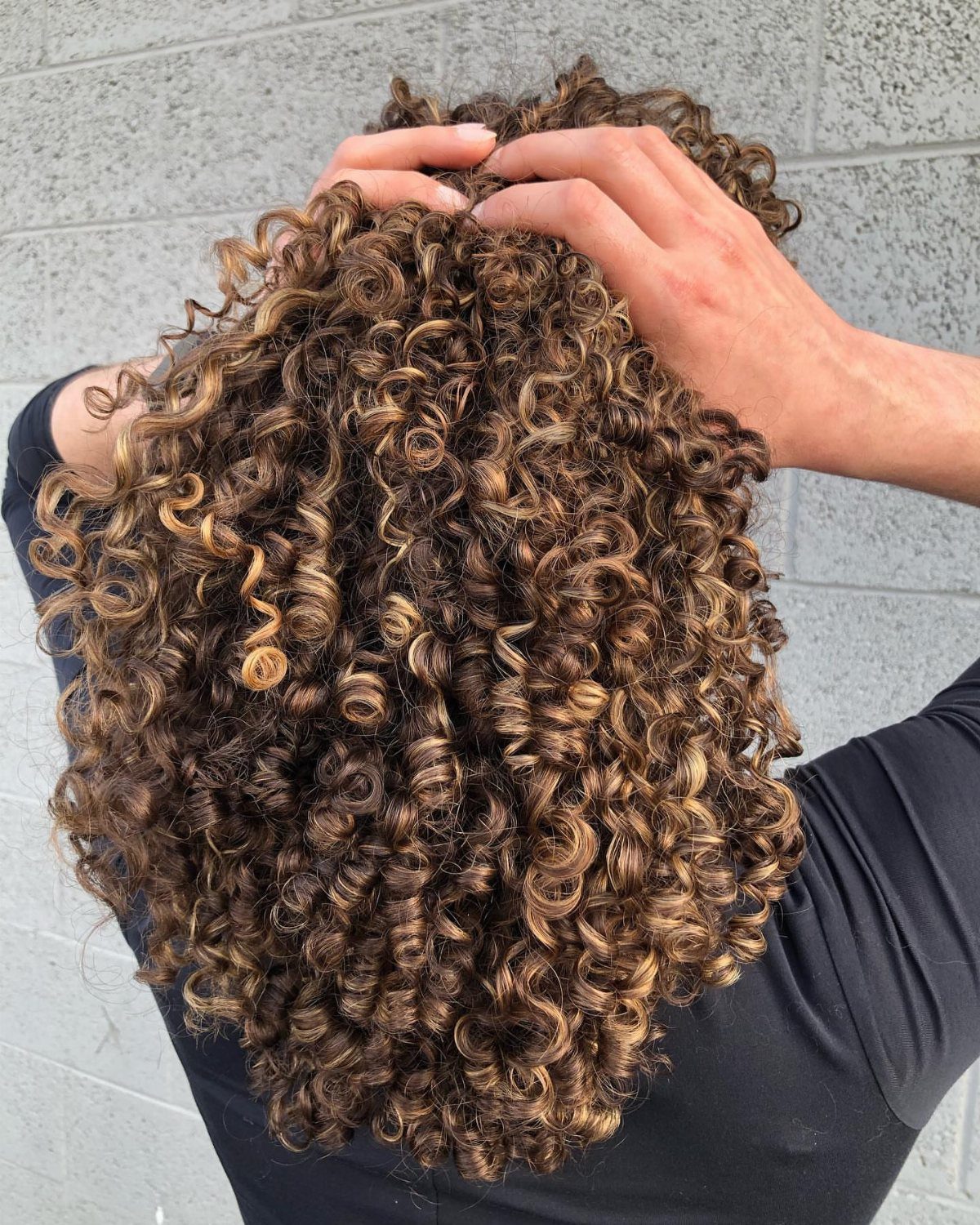 Short highlighted curls lifted by hands against a light gray block wall