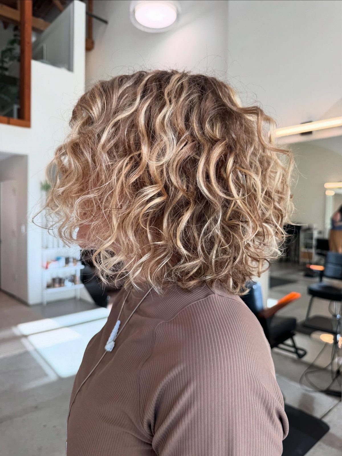 Layered curly lob with blonde-highlighted ringlets seen from behind in a salon