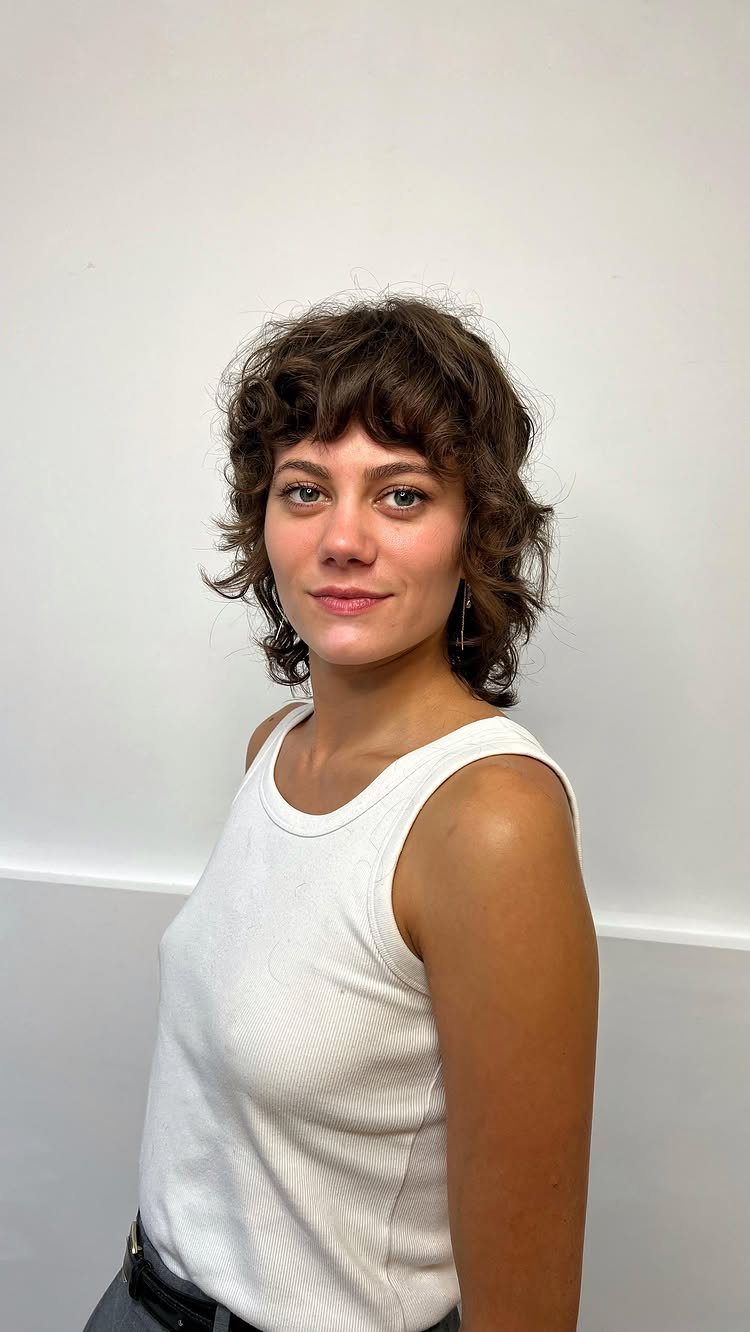 Woman with short layered brown curls and tousled bangs wearing a white sleeveless top