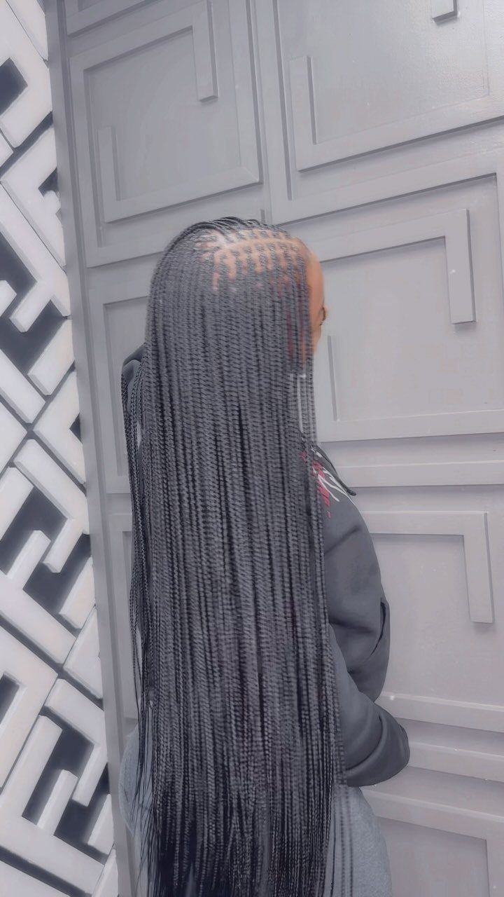 Long fine black braids fall down the back against a gray paneled wall