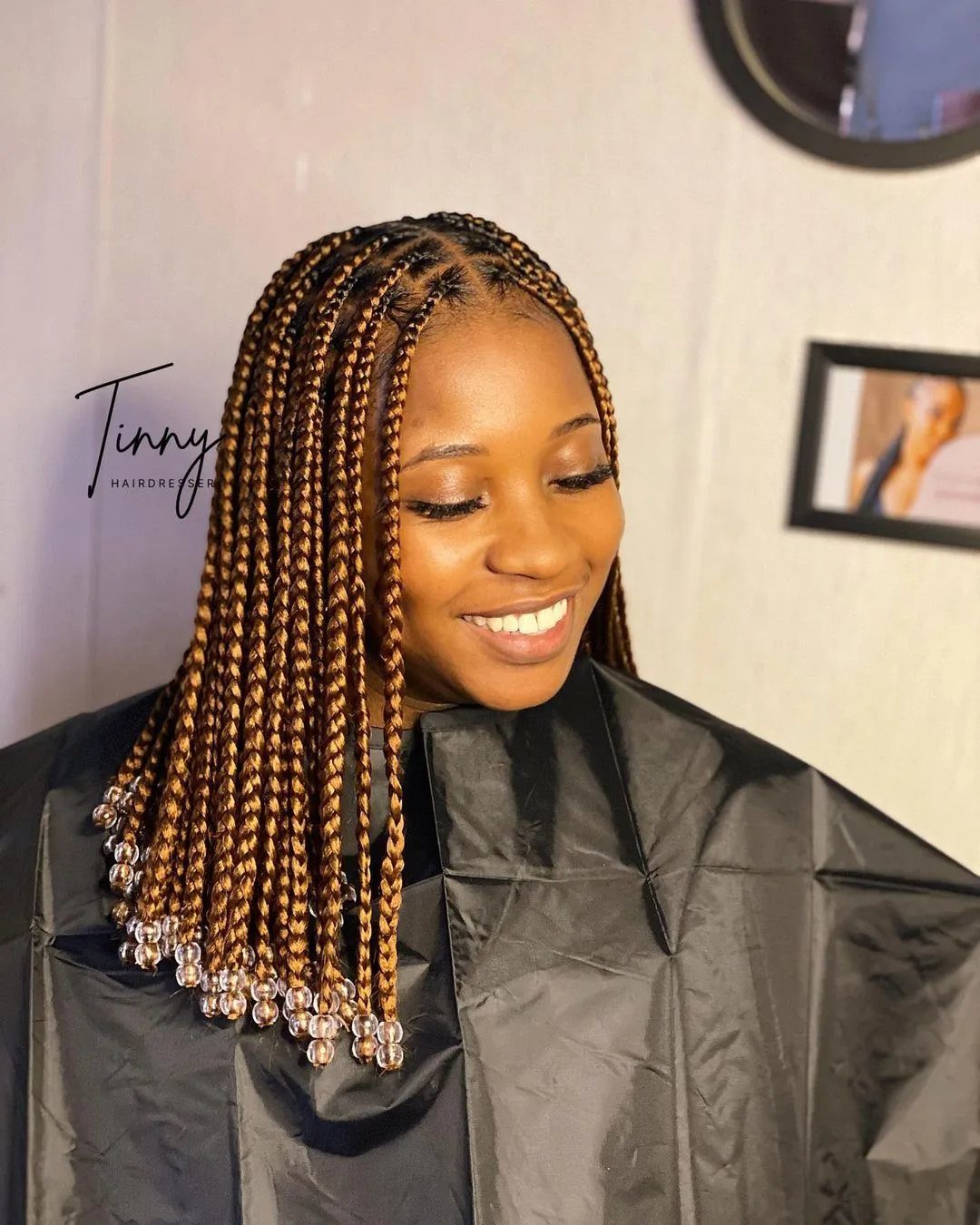 Long small braids ending in clear beads drape over a smiling woman’s salon cape