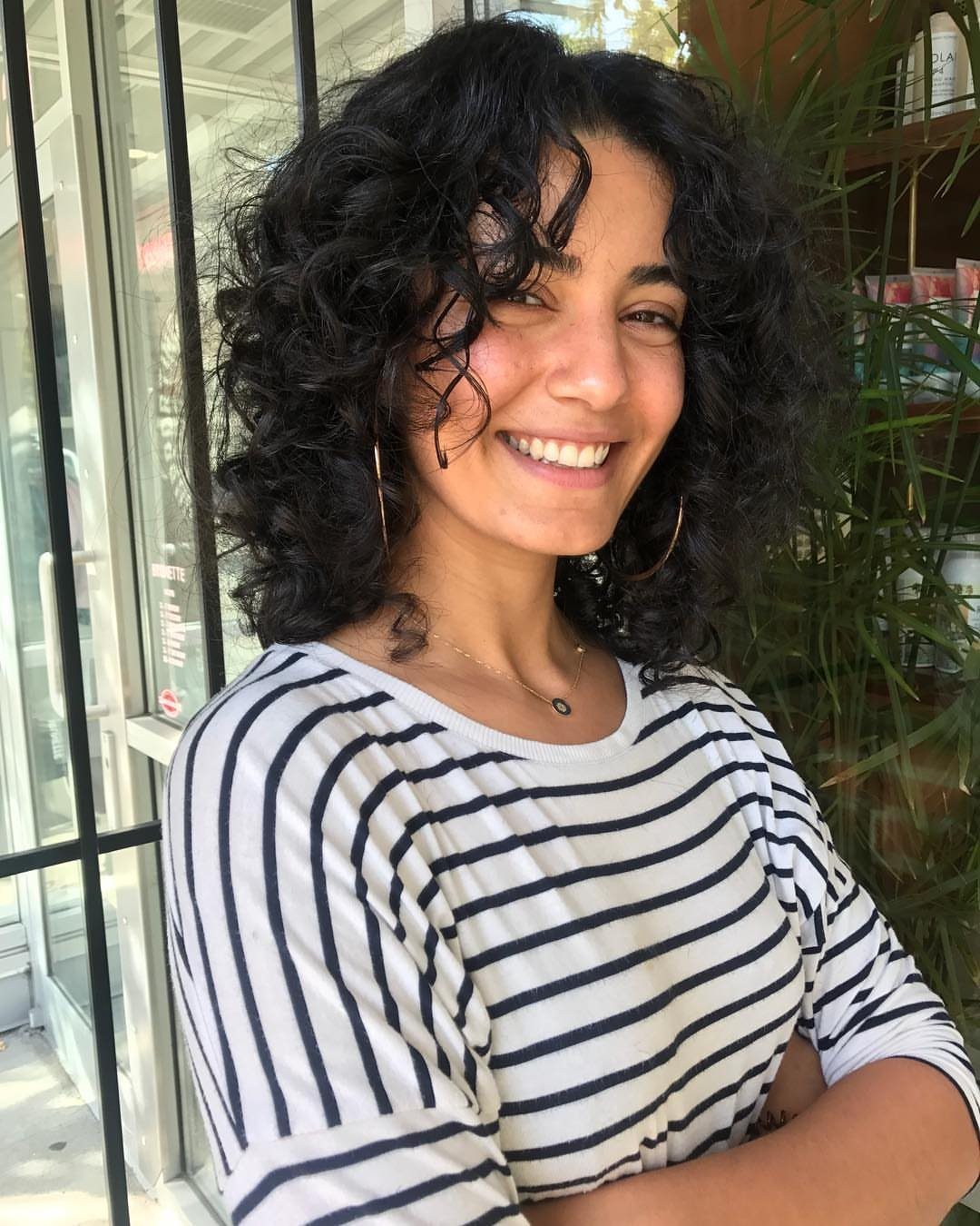 Woman showing shoulder-length layered curly hair beside windows and leafy plants