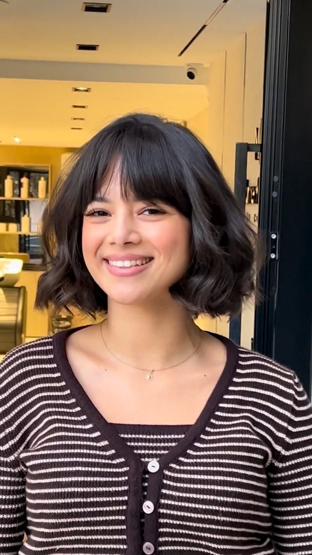 Woman smiling with a short wavy dark bob and forehead bangs in a salon